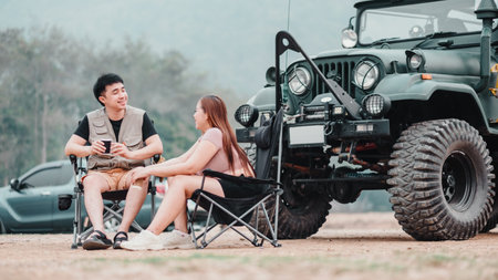 Couple is deep in conversation, enjoying a warm drink while seated beside a rugged off-road car at their scenic campsite.の写真素材