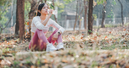 Fitness conscious woman drinking water while resting after a workout session in a sunlit forest...