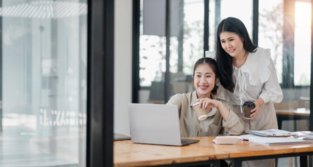 Two smiling female colleagues collaboratively working on a project with a laptop and coffee in a bright office space.の写真素材