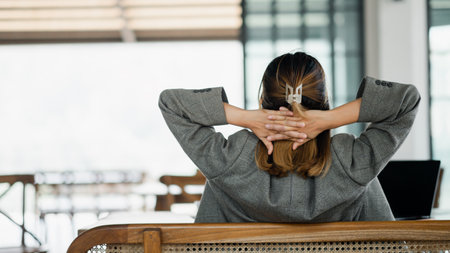 Rear view of a businesswoman taking a break at her workspace, with hands clasped behind her head and laptop on the table.の写真素材