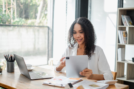 Cheerful businesswoman reviews a document with her laptop open, seated at her desk in a well-lit...