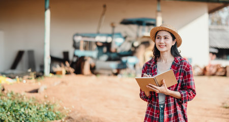 Optimistic young farmer holding a notepad stands in front of farm equipment, ready to tackle the day's tasks.の写真素材
