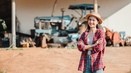 Confident female farmer stands with crossed arms in front of agricultural machinery on a sunny rural...