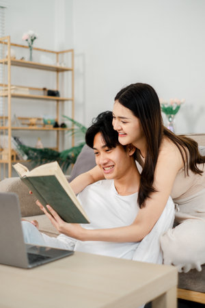 young man in a white t-shirt and a woman in a beige sleeveless dress are engaged in reading a hardcover book together on a sofa. A laptop is open beside them.の写真素材