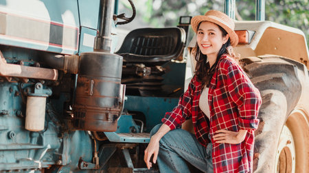 Joyful farmer in a straw hat and plaid shirt leans on a blue tractor, posing with hands on her hips.の写真素材