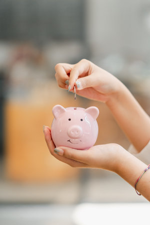 Hands of a person inserting a coin into a smiling pink piggy bank, symbolizing savings and financial planning.の写真素材