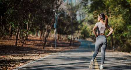 A woman is running on a road with trees in the background. She is wearing a grey shirt and grey...