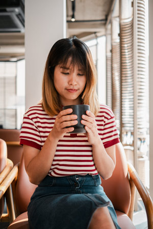 A woman in a striped shirt is sitting in a chair and holding a cup. She is in a relaxed and...