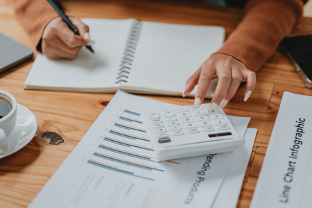 A woman is writing in a notebook with a calculator on the table. The notebook has a line chart on it