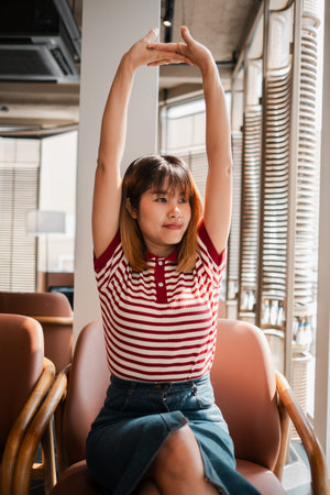 A woman in a red and white striped shirt is stretching her arms out in front of her. She is sitting in a chair with her legs crossed. Concept of relaxation and comfortの写真素材