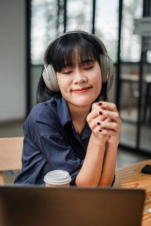 A woman wearing headphones is sitting at a table with a laptop and a cup of coffee. She is smiling...