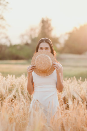 A woman is standing in a field of tall grass, holding a straw hat over her face. The scene is serene...
