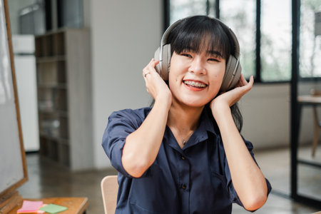 A woman wearing headphones and smiling. She is wearing a blue shirt. The room has a white board on the wallの写真素材