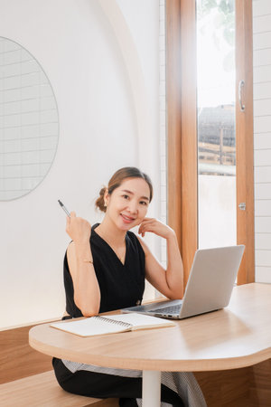 A woman is sitting at a table with a laptop and a pen. She is smiling and she is enjoying her work