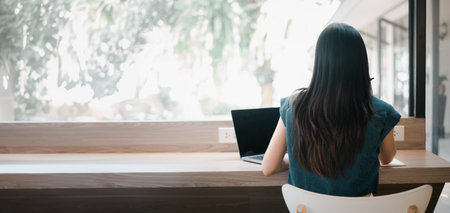 A woman sits at a desk with a laptop in front of her. She is wearing a blue shirt and has long hair....