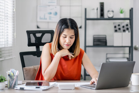 A woman in an orange shirt is sitting at a desk with a laptop and a tablet. She is looking at the laptop screen and she is focused on her workの写真素材