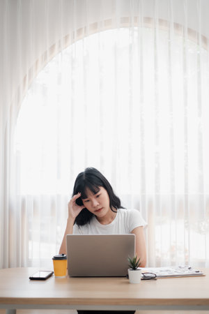 A young woman working on her laptop at a home office desk with coffee and documents in a bright room with a large window and sheer curtains.の写真素材
