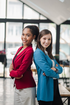 Two confident businesswomen standing back to back in a modern office with large windows, showcasing teamwork and professionalism.の写真素材