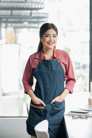 Smiling female barista in a denim apron standing in a modern cafe, ready to serve customers. Bright and airy coffee shop interior.の写真素材