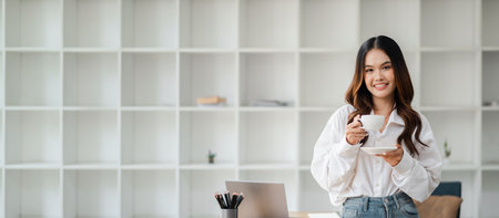 Smiling young woman holding a cup of coffee in a modern office setting with minimalist white shelving in the background.の写真素材