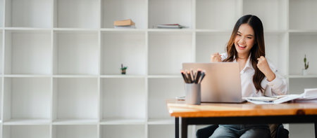 Excited young woman celebrating success while working on a laptop in a modern office with minimalist white shelves in the background.の写真素材