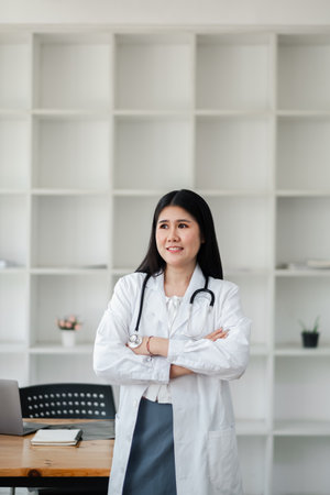A confident female doctor in a white coat with a stethoscope standing in a modern office with shelves in the background.の写真素材