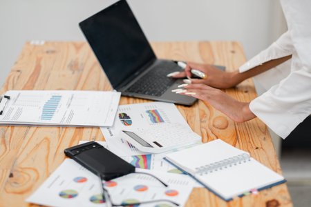 Professional businesswoman analyzing financial data on a laptop with various documents and charts on...