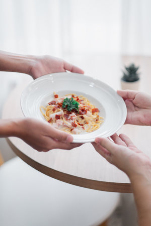 Close-up of hands passing a plate of pasta with bacon and parsley in a bright, modern dining setting.の写真素材