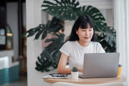 Asian woman working on a laptop at home office with coffee and plant, showcasing a modern and bright workspace.の写真素材
