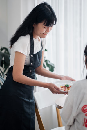 A woman in a black apron serves a dish to a customer in a bright, modern cafe with white interiors...