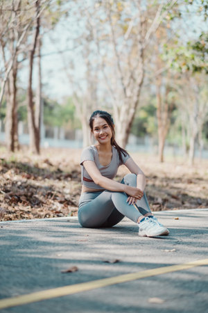 Smiling young woman in athletic wear sitting on a park pathway surrounded by trees, enjoying a sunny day.の写真素材