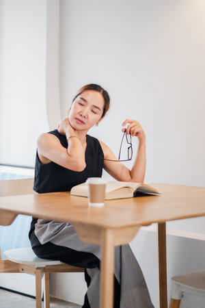 A woman in a modern cafe, holding glasses, stretching her neck, and relaxing with a book. Casual and comfortable setting.の写真素材