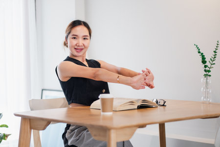Woman stretching at desk with coffee and book in a modern home office. Bright and minimalist workspace with natural light and greenery.の写真素材