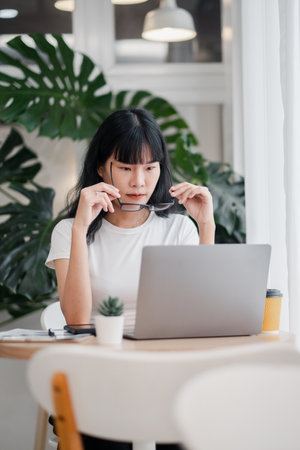 Focused young woman in casual attire working on a laptop at a home office with a tropical plant background.の写真素材