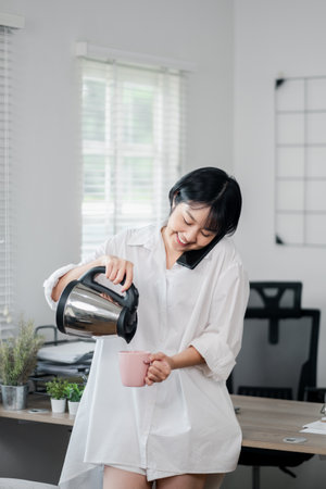A young woman in a white shirt pouring coffee into a pink mug in a modern home office with natural light and plants.の写真素材