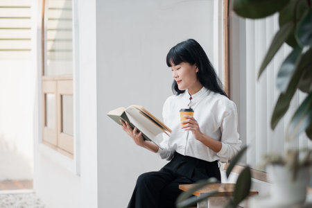 A young woman enjoys a peaceful moment reading a book and sipping coffee in a bright, modern...