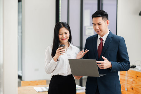 Two business professionals in formal attire discussing work while using a laptop in a modern office setting.の写真素材