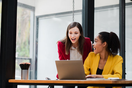 Two businesswomen in vibrant blazers working together on a laptop, showcasing teamwork and collaboration in a contemporary office environment.の写真素材