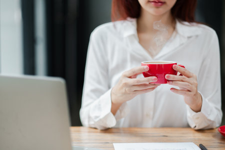 A woman in a white shirt holds a red coffee mug while working on a laptop at a wooden desk in a modern office setting.の写真素材