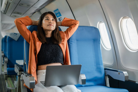 A young woman relaxes in an airplane seat with her laptop, enjoying a comfortable flight in a modern aircraft cabin.の写真素材
