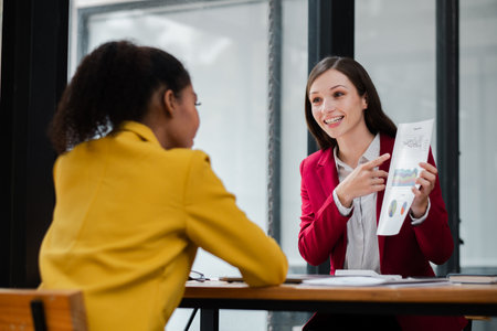 Two professional women engaged in a business meeting, discussing documents in a modern office environment with large windows.の写真素材