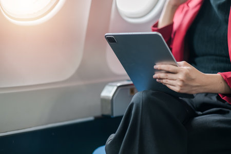 A businesswoman in a red blazer using a tablet while seated on an airplane, showcasing modern travel and technology.の写真素材