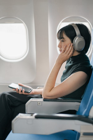 A young woman with headphones, seated on an airplane, listening to music and holding a smartphone,...