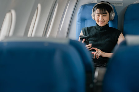 A woman wearing headphones and using a smartphone while seated on an airplane, enjoying in-flight...
