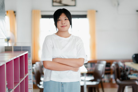A young woman stands confidently in a bright, modern classroom with her arms crossed, exuding self-assurance and determination.の写真素材