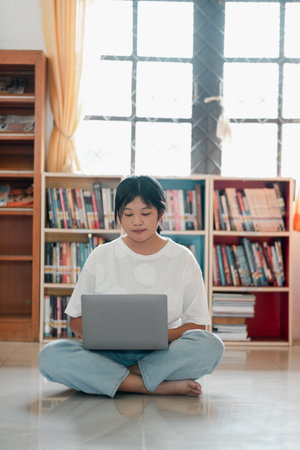 A young woman sits cross-legged on the floor, working on her laptop in a bright home library filled...