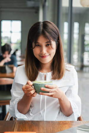 A young woman in a white blouse smiles while holding a cup of coffee in a bright, modern cafe with...