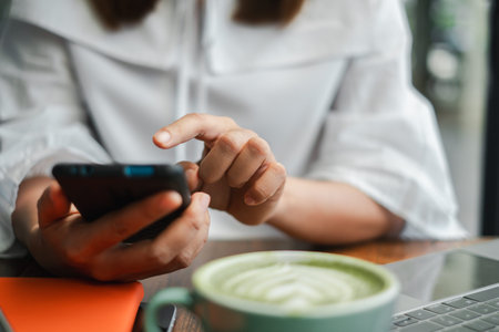 Close-up of a woman using a smartphone in a cozy cafe, with a cup of matcha latte and a laptop on...