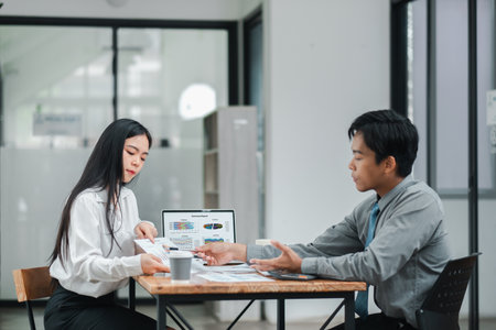 Two business professionals in a modern office setting, engaged in a discussion with documents and a laptop displaying data.の写真素材