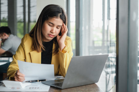 Young woman in a yellow blazer working on a laptop, appearing stressed while reviewing documents at a home office.の写真素材